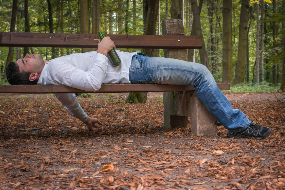 depressed drunk man asleep outdoor on a park bench with beer bottles