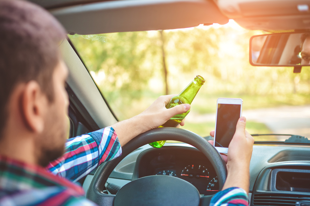 Drunk young man driving a car with a bottle of beer and mobile phone. Don't drink and drive
